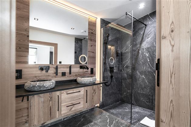 Een moderne badkamer met houten panelen en elegante zwarte tegels. De douche is afgescheiden door een glazen wand en er zijn twee wastafels van natuursteen.