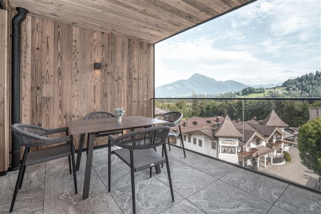 Een modern ingericht balkon met een houten vloer en een houten tafel. Op de achtergrond zijn bergen en een idyllisch stadslandschap te zien.