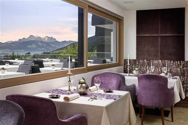 An elegant restaurant with purple chairs and table decoration. In the background, majestic mountains and a beautiful sky are visible.