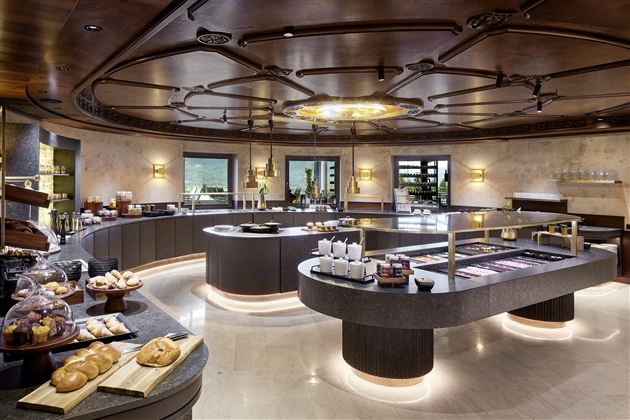 An elegant dining room with a round buffet and appealing lighting. On the tables, fresh pastries and various dishes are arranged.