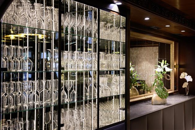 An elegant glass shelf with many glasses in a stylish bar. In the background, plants and a cozy decor can be seen.