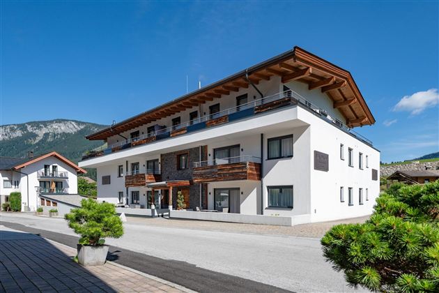 Ein modernes Gebäude mit mehreren Wohnungen, umgeben von Bäumen und einer klaren blauen Himmel. Im Hintergrund sind sanfte Berge zu sehen.