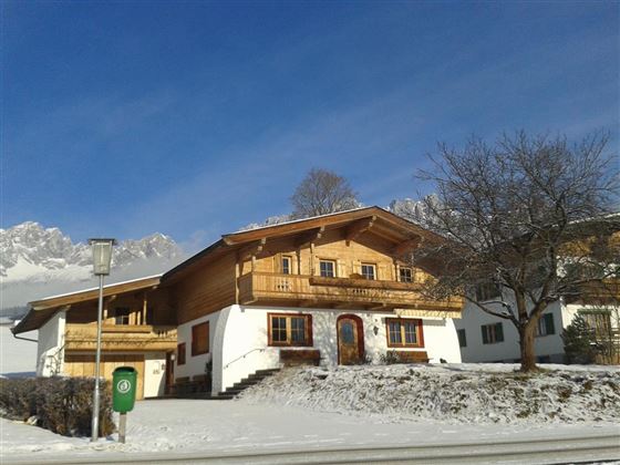 A typical alpine house with wooden cladding in a snowy environment. In the background, the mountains and a clear sky can be seen.