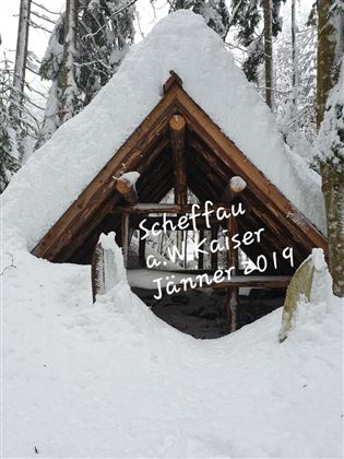A small wooden cabin, covered in snow, surrounded by snow-covered trees. The atmosphere is calm and wintry.