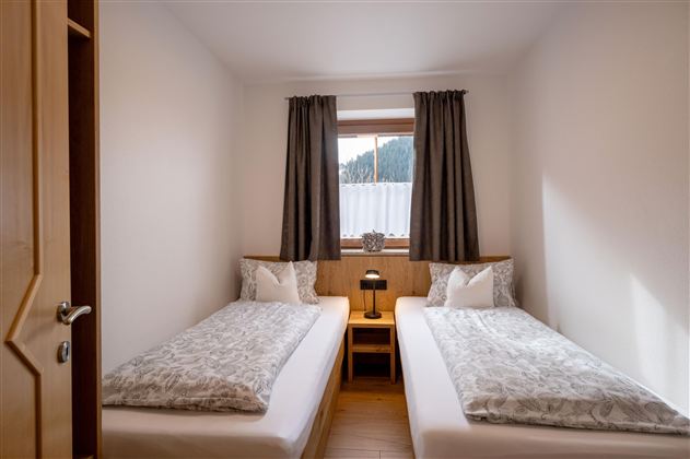 Een lichte slaapkamer met twee eenpersoonsbedden en een klein nachtkastje. Voor het raam zijn er ondoorzichtige gordijnen en een mooi uitzicht op de bergen.