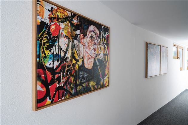 Ein farbenfrohes Kunstwerk hängt an einer weißen Wand im Flur. Im Hintergrund sind weitere Bilder und ein Fenster sichtbar.