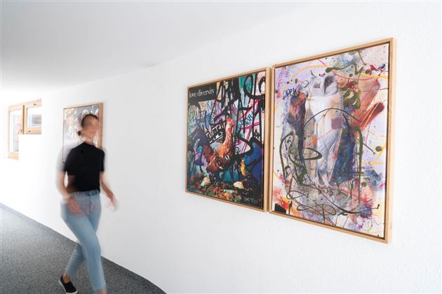 Eine Person geht an bunten Gemälden entlang, die an einer weißen Wand hängen. Der Raum hat ein modernes und helles Ambiente.