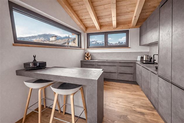 Eine moderne Küche mit Holzdecken und großen Fenstern. Der Raum ist mit grauen Möbeln und einem Tisch mit Hockern ausgestattet.