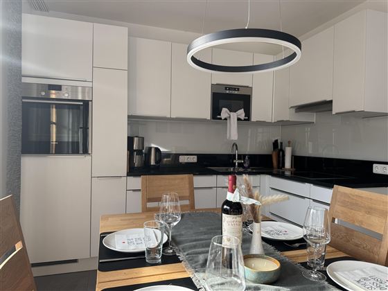 Moderne keuken met elegante witte kasten en een houten tafel die gedekt is. Op de achtergrond zijn een koelkast en een fornuis zichtbaar, evenals decoratieve elementen.