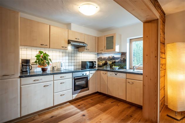 A modern kitchen with wooden cabinets and a large window. In the foreground, there is a plant and a refrigerator is visible.