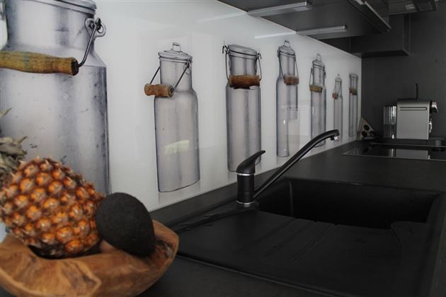 Eine moderne Küche mit einem schwarzen Waschbecken und einer Wandverkleidung, die stilisierte Gläser zeigt. Auf der Arbeitsfläche befinden sich eine Ananas und ein schwarzer Gegenstand in einer Holzschale.