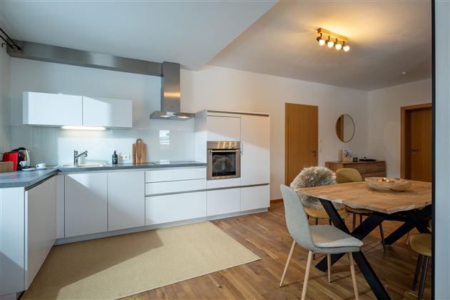 Eine moderne Küche mit weißen Schränken und Holzfußboden. Der Essbereich ist hell und einladend mit einem großen Tisch und Stühlen.