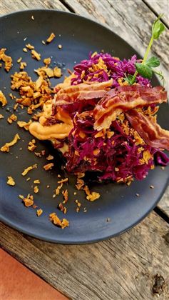 Een bord met paarse coleslaw, knapperige spekjes en gebakken uien. Het gerecht ligt op een zwart bord en is decoratief opgediend.
