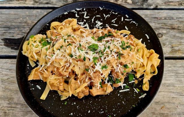 Een bord met pasta en een romige saus, gegarneerd met verse Parmezaanse kaas en peterselie. De pasta is op een zwart bord geserveerd en staat op een rustieke houten ondergrond.