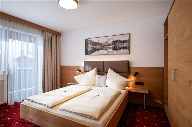 Ein gemütliches Schlafzimmer mit einem großen Bett, dekorierten Kissen und Holzmöbeln. Die großen Fenster lassen viel Tageslicht herein.