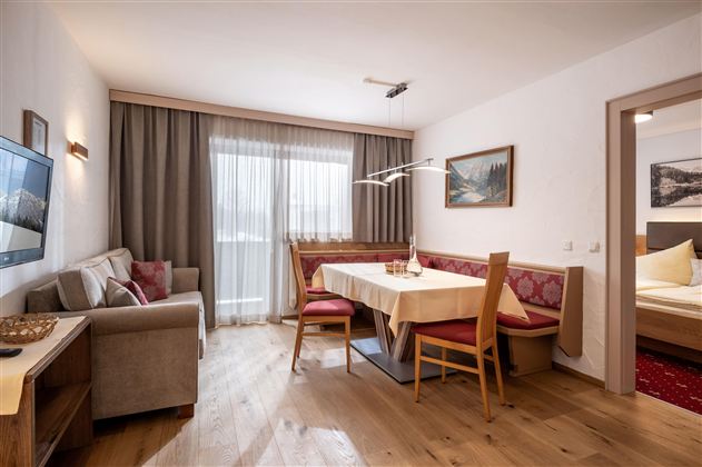 Ein gemütlicher Wohnraum mit einem Tisch und Stühlen. Durch ein großes Fenster strömt Tageslicht herein.