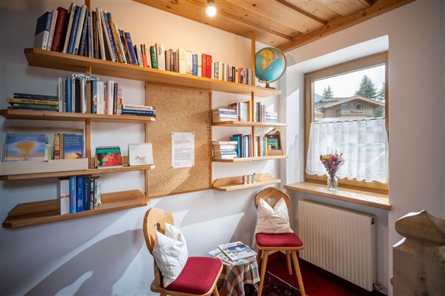 A cozy reading nook with a bookshelf on the wall. Two chairs and a small table are positioned in front of a window with a view outside.