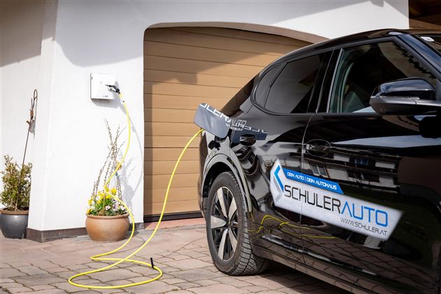 A black electric car is being charged at a charging station. In the background, a garage can be seen.