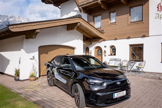 A black car is parked in front of a traditional, wooden-clad house. In the background, some garden furniture and a clear sky are visible.