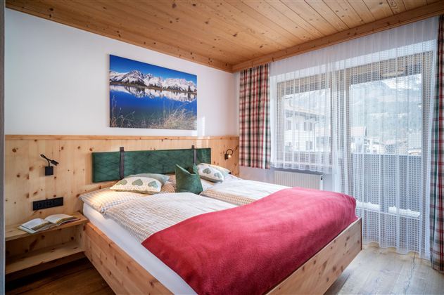A cozy bedroom with a large bed and wooden decor. The room has a balcony and a beautiful picture of mountains on the wall.