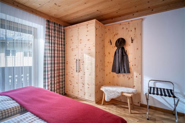 A cozy bedroom with wooden walls and a large wardrobe. On a bed lies a red blanket and beside it stands a bench with a hat.