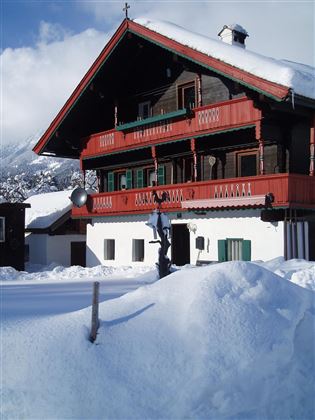 Ein traditionelles Holzhaus mit roter Balkonverzierung, umgeben von Schnee. Im Hintergrund sind Berge und ein klarer Himmel zu sehen.