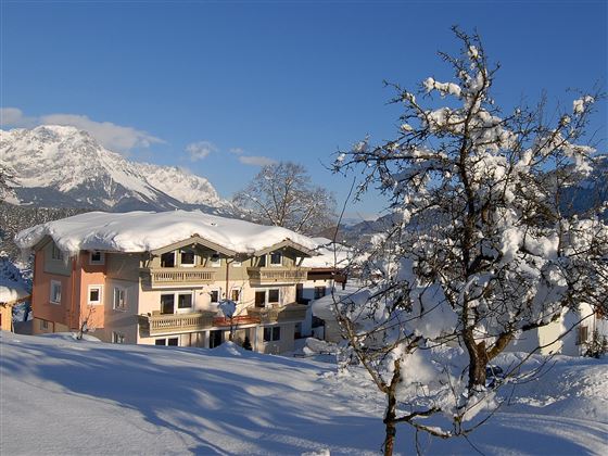 Ein gemütliches Haus im Schnee mit einem strahlend blauen Himmel. Die Umgebung ist von schneebedeckten Bergen und Bäumen geprägt.