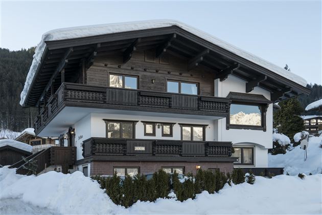 An impressive chalet in the Alps, surrounded by snow. The building has a traditional wooden style with balconies and large windows.
