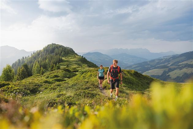 Zwei Wanderer steigen auf einem berghängigen Pfad. Im Hintergrund sind sanfte Hügel und eine beeindruckende Landschaft zu sehen.