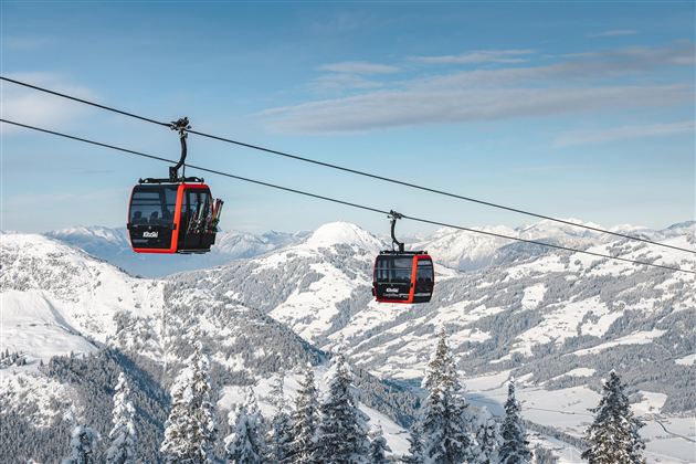 Eine Seilbahn fährt über schneebedeckte Berge. Die Landschaft ist geprägt von winterlichen Tannen und weitem Himmel.
