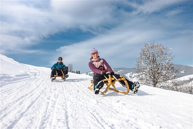 Zwei Personen rodeln auf einem schneebedeckten Hang. Der Himmel ist klar und blau, und die Landschaft ist winterlich schön.