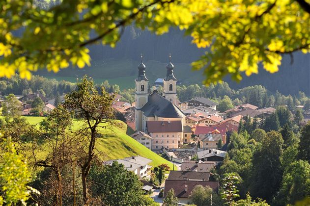 Ein malerisches Dorf in einer grünen Umgebung mit einer Kirche im Vordergrund. Die sanften Hügel und Bäume verleihen der Szene eine ruhige Atmosphäre.