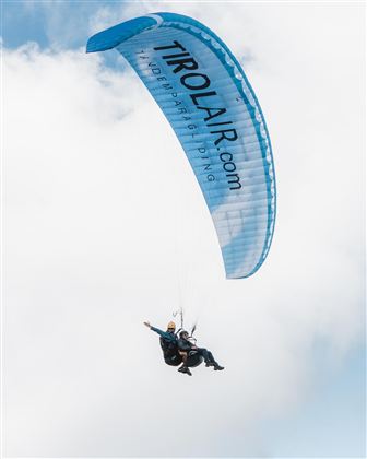 Ein Tandem-Gleitschirmflieger schwebt durch den Himmel. Der Gleitschirm hat eine blaue Farbe mit der Aufschrift "Tirol Air".