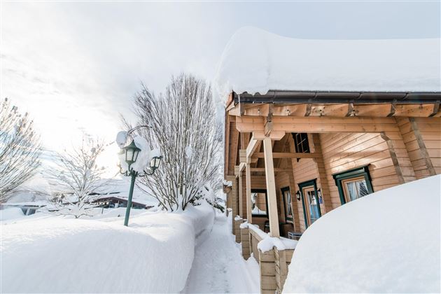 Ein Holzhaus unter einem großen Schneedecken, umgeben von schneebedeckten Bäumen. Die Sonne scheint durch die Wolken und sorgt für eine wintry, ruhige Atmosphäre.