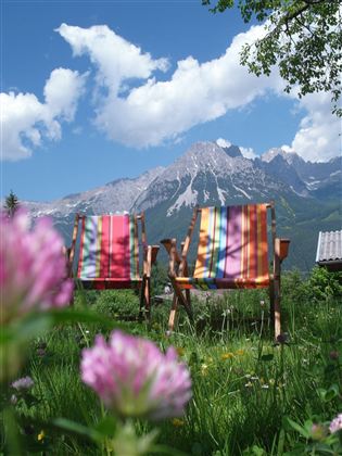 Zwei bunte Liegestühle stehen in einer grünen Wiese mit Blumen. Im Hintergrund sind majestätische Berge und ein blauer Himmel mit einigen Wolken zu sehen.