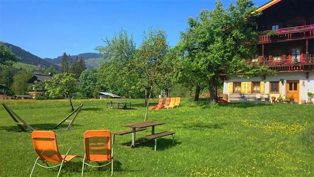 Een groene wei met comfortabele ligstoelen en schaduwgevende bomen. Op de achtergrond staat een gebouw met balkons en uitzicht op de bergen.