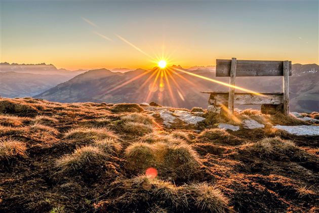 A beautiful sunrise over the mountains. A wooden bench stands in the foreground on the grass.