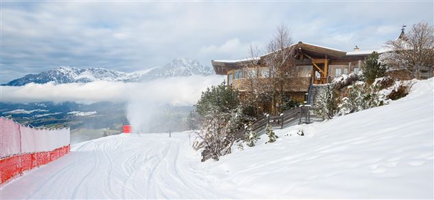 Eine verschneite Landschaft mit einem modernen Gebäude und schneebedeckten Bergen im Hintergrund. Am Fuße des Berges ist eine Skipiste zu sehen.