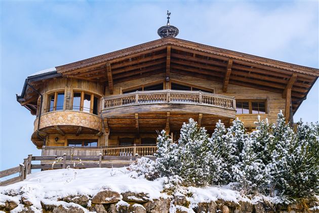 Ein gemütliches Holzhaus mit Balkon und großen Fenstern. Es liegt in einer verschneiten Landschaft mit Tannenbäumen.