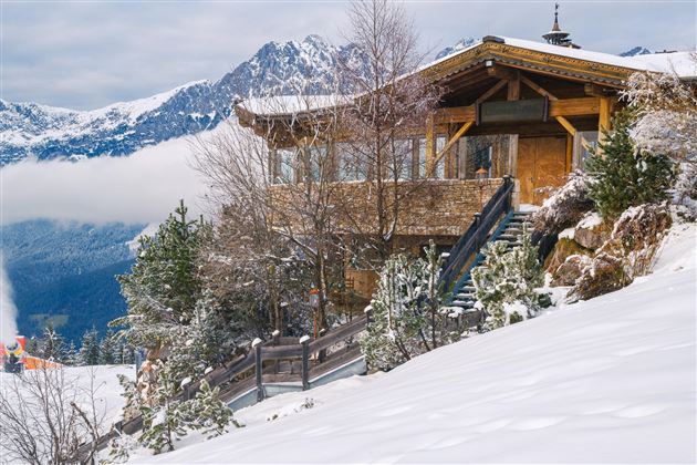 Ein gemütliches Holzhaus im Schnee, umgeben von Bäumen und Bergen. Der Himmel ist bewölkt und die Landschaft wirkt friedlich.