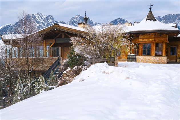 Ein gemütliches Holzhaus in einer verschneiten Landschaft. Im Hintergrund sind beeindruckende Berge zu sehen.