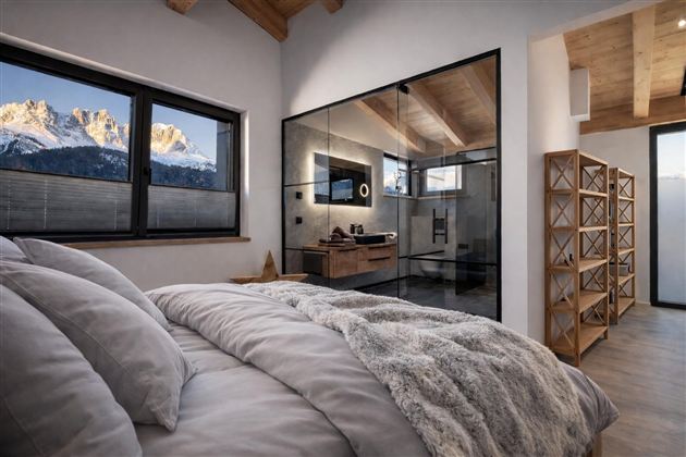 Ein modernes Schlafzimmer mit einem komfortablen Bett und einem Blick auf schneebedeckte Berge. Das Bad ist durch eine Glaswand getrennt und strahlt Eleganz aus.