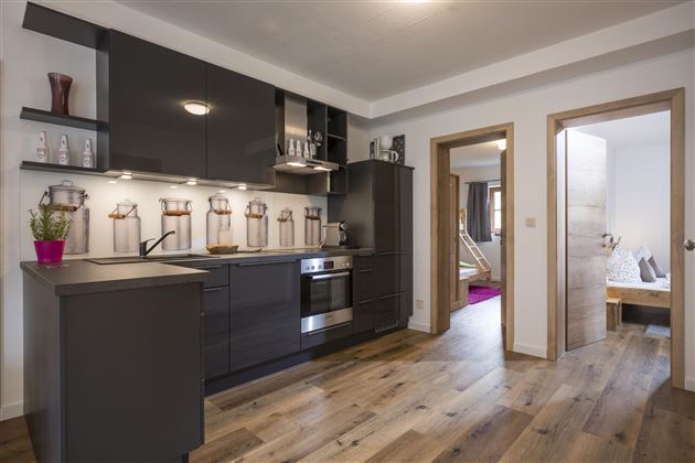 Eine moderne Küche mit dunklen Schränken und hellen Arbeitsflächen. Der Raum ist hell und einladend, mit Holzfußboden und einem offenen Durchgang zu einem Wohnbereich.