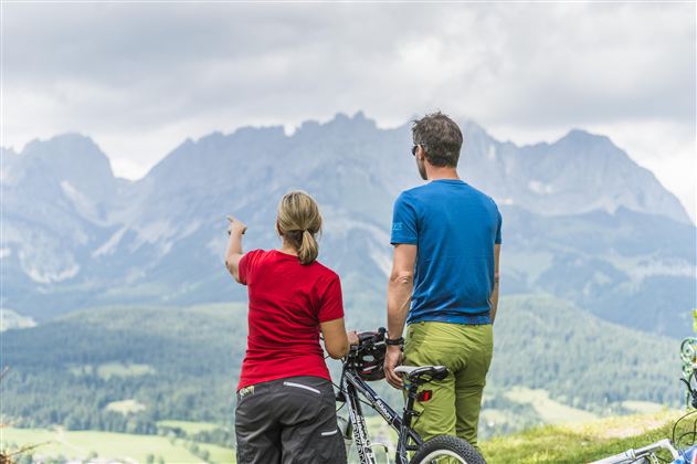 A couple is standing on a hill and looking at the mountains. They have bicycles with them and are enjoying the view.