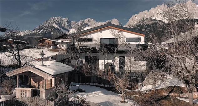 Ein modernes Haus in einer schneebedeckten Landschaft mit Bergen im Hintergrund. In der Nähe steht eine traditionelle Holzhütte, umgeben von Bäumen.