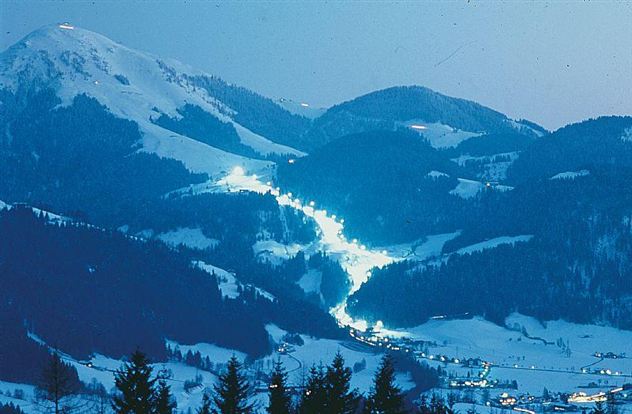 Eine winterliche Berglandschaft mit schneebedeckten Hügeln. Sanfte Lichter erleuchten die Piste und das Dorf im Tal.