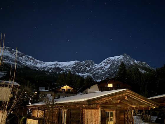 Een schilderachtig berglandschap bij nacht met met sneeuw bedekte toppen en een heldere sterrenhemel. Op de voorgrond is een rustiek houten huis te zien.