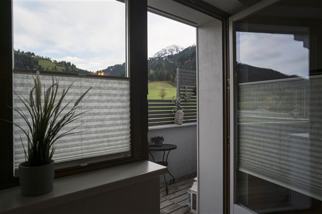 Een blik op een terras met een plant in een pot en mooie berglandschappen op de achtergrond. De ramen tonen een rustige landschap met zachte heuvels.