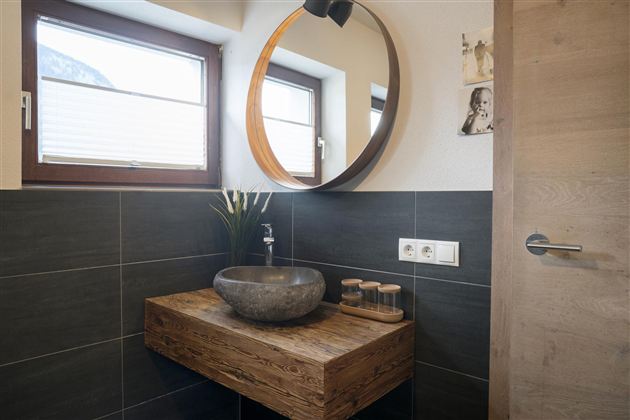 Een moderne badkamer met een grote ronde spiegel en een stijlvolle wastafel van steen. De muren zijn in donkere tegels uitgevoerd, en er zijn enkele decoratieve elementen zoals planten.