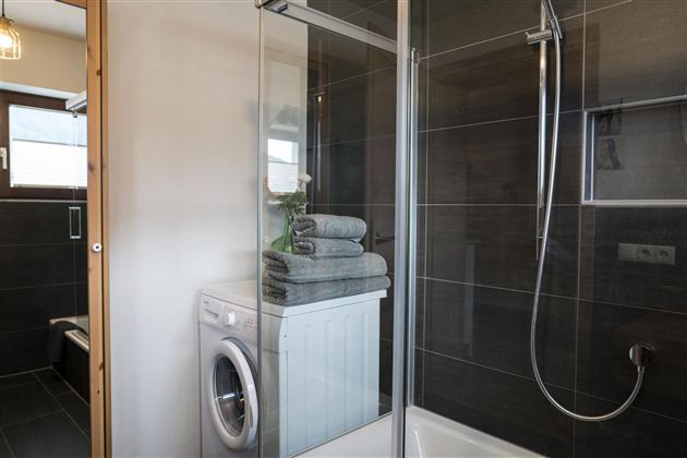 Een moderne badkamer met een douche en een wasmachine. Stapels grijze handdoeken liggen op de wasmachine.